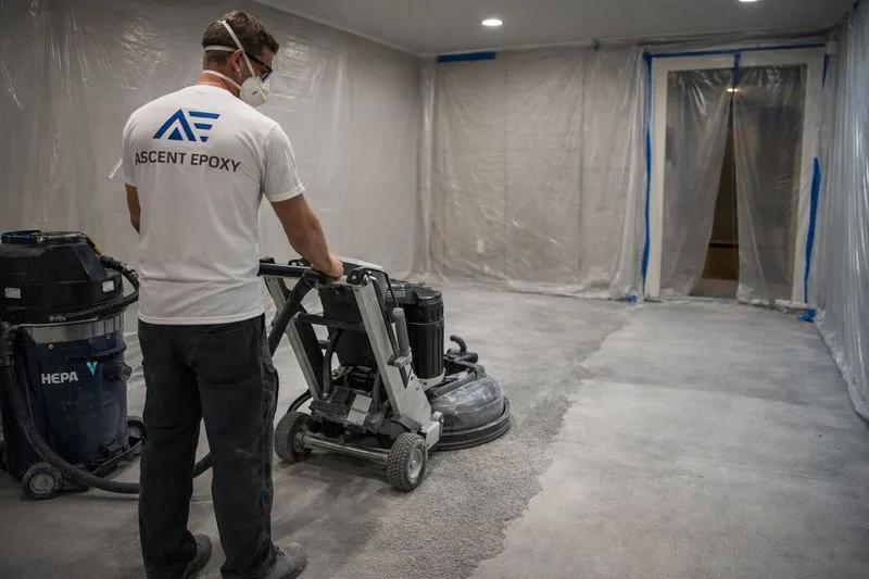 Ascent Epoxy installer operating diamond grinder on residential basement concrete with HEPA dust extraction and plastic sheeting protecting walls